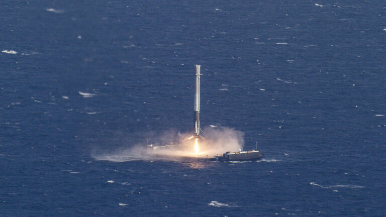 A rocket booster lands on a platform in the ocean, with flames visible at its base upon landing. The platform is floating on calm, blue waters.