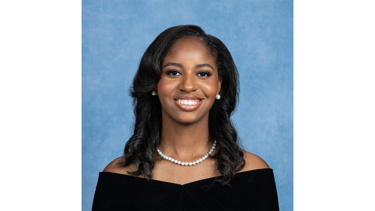 A woman with long dark hair wearing a black off-the-shoulder top and a pearl necklace smiles in front of a blue background.