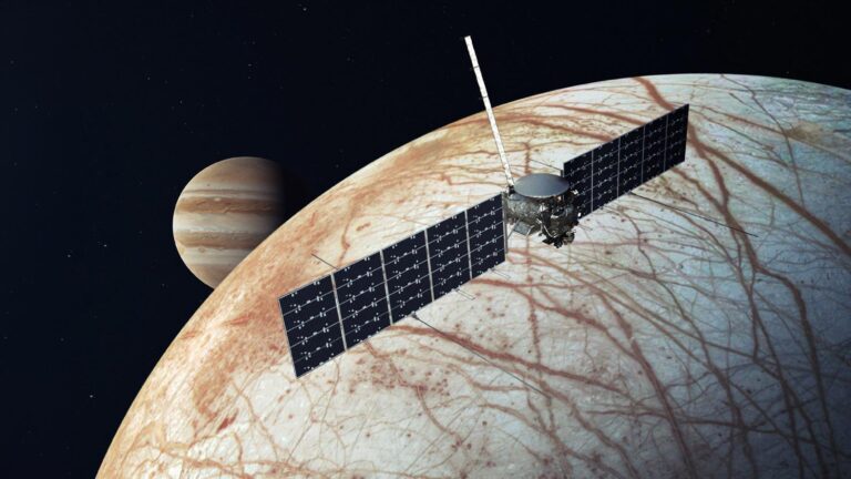 A spacecraft with large solar panels orbits a moon with Jupiter in the background. The moon's surface appears cracked and icy.