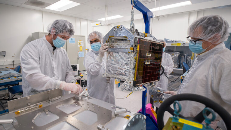 Scientists in a lab wearing protective gear and masks work on a metallic, foil-covered satellite or spacecraft component.