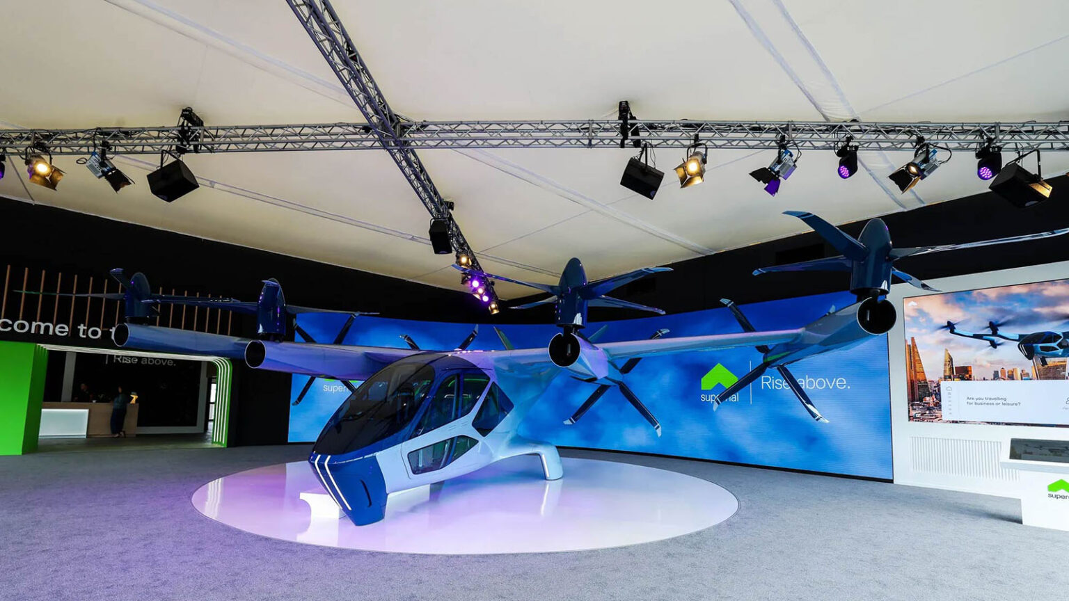 A blue, futuristic aircraft with multiple propellers is displayed in an indoor exhibition area with overhead lighting and a large screen in the background.