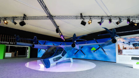 A blue, futuristic aircraft with multiple propellers is displayed in an indoor exhibition area with overhead lighting and a large screen in the background.
