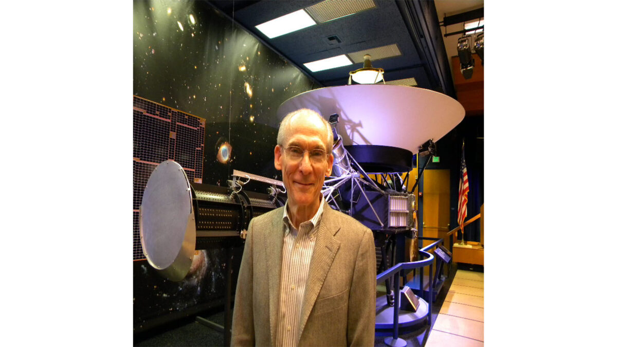 An older man in a suit stands in front of a large spacecraft model displayed in a room with an American flag and cosmic background.