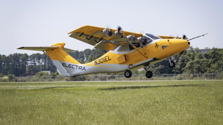 A yellow and white Electra aircraft with visible registration number N301EL takes off from a grassy runway. Trees are in the background under a clear sky.