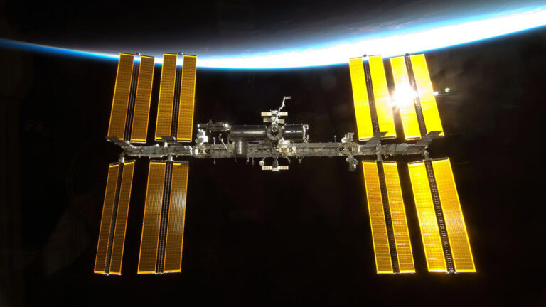 The International Space Station floats in space with its solar panels extended, illuminated by sunlight, with Earth visible in the background.