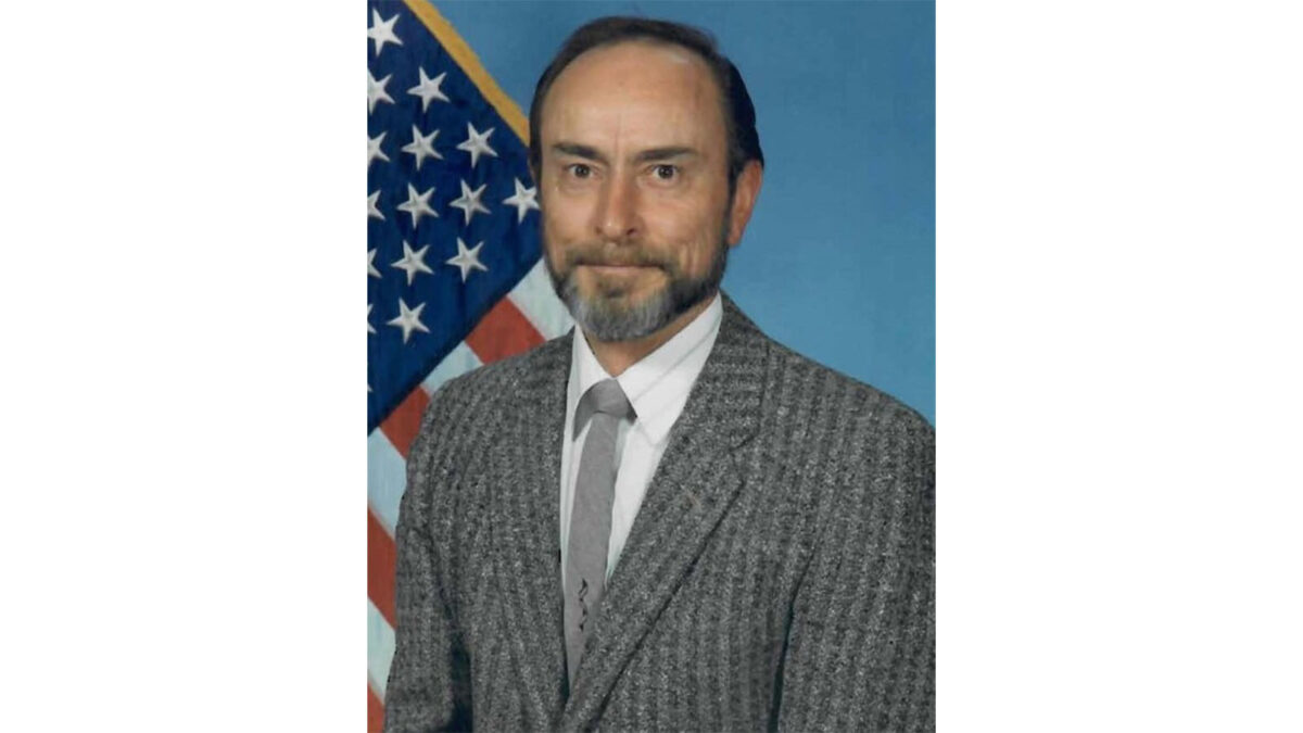 A middle-aged man with a beard and mustache wearing a grey suit poses in front of an American flag and blue background.