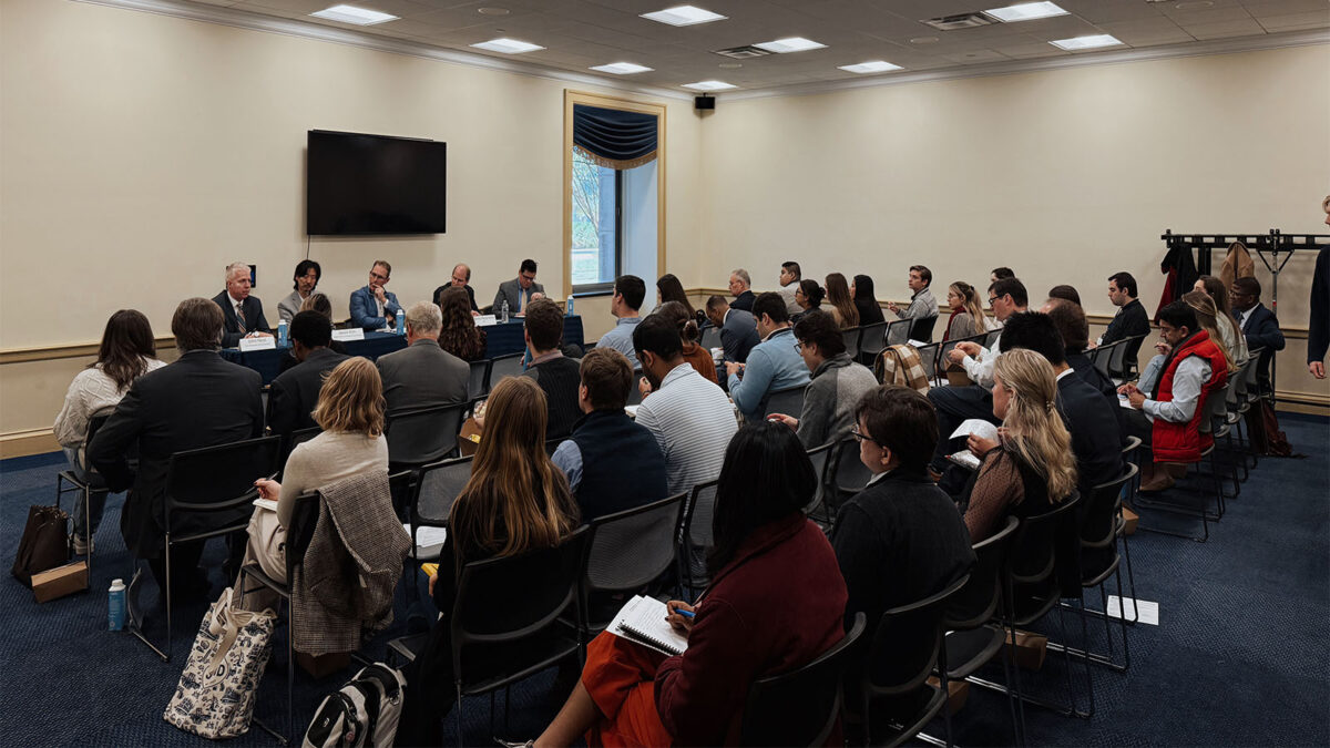 A group of people are seated and listening to a panel of speakers in a conference room. A screen is mounted on the wall behind the speakers.