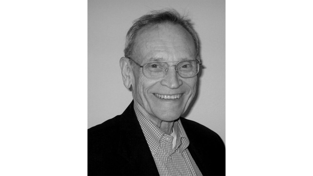 An elderly man with glasses and a smile, wearing a suit jacket and checkered shirt, in a black and white portrait.