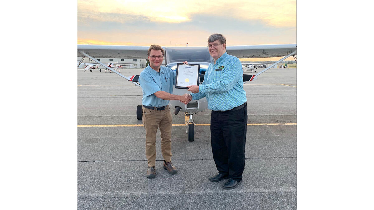 Two men shaking hands in front of a small plane. One holds a framed certificate. Both are wearing blue shirts and glasses.