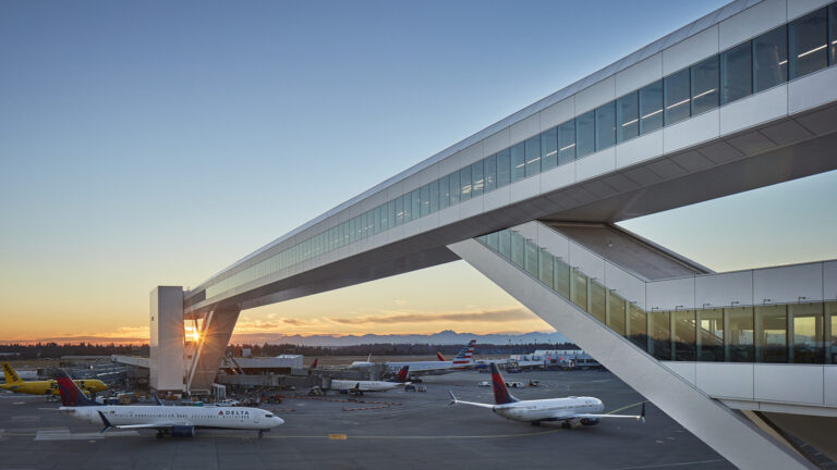 Airport scene at sunset featuring parked airplanes, a skybridge, and a terminal building with mountains in the background.