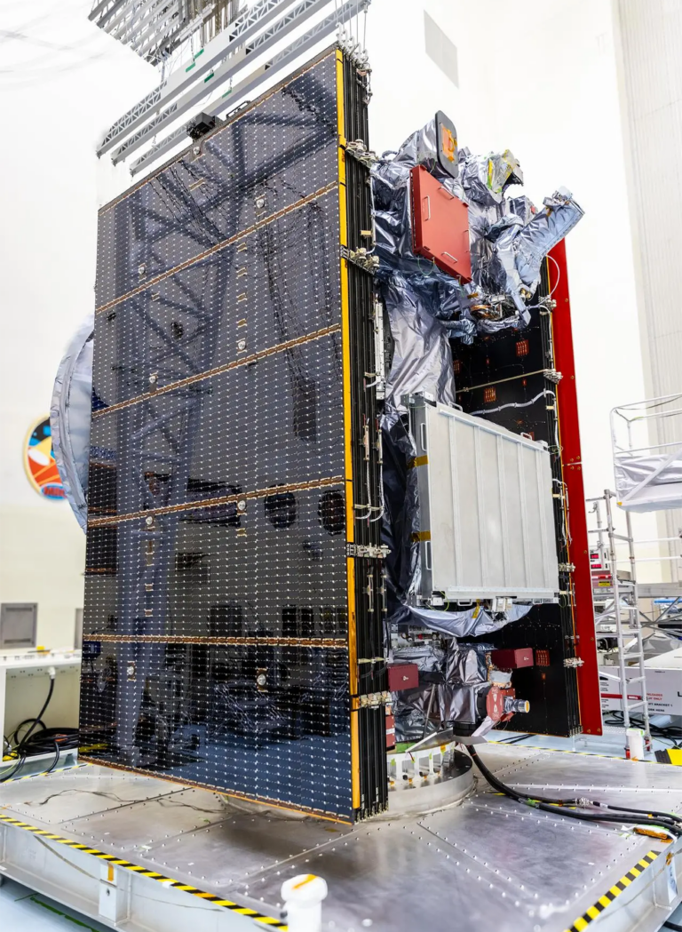 A large satellite with visible solar panels and various electronic components is positioned upright in an indoor facility.