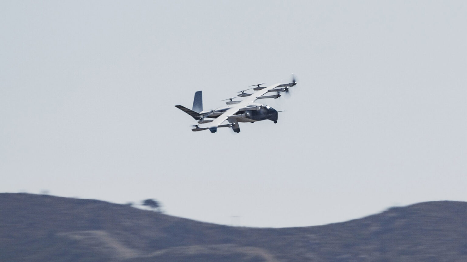 Flying vehicle with multiple propellers in the sky above distant hills.