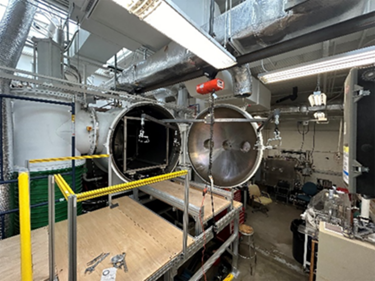 A large industrial chamber with open circular doors, surrounded by metal pipes and equipment, in a spacious room with exposed ceilings.