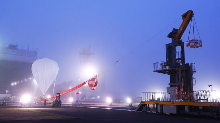 Scientific balloons reach new heights A large white balloon tethered to a crane in a foggy, illuminated area with machinery and workers nearby.