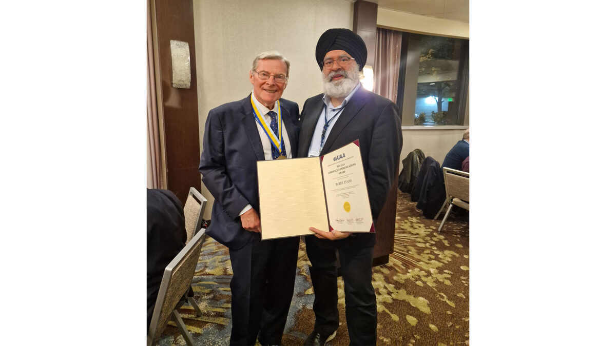 Two men in suits stand together in a room, smiling, with one holding a large certificate.