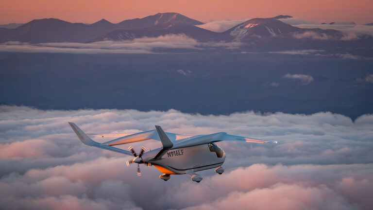 A small airplane flies above clouds during sunset, with distant mountains visible in the background.