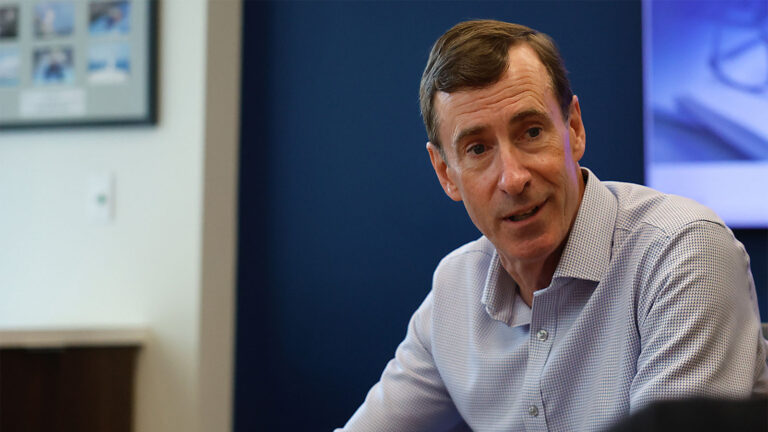 Man in a collared shirt sitting at a table, engaging in conversation. A framed picture is visible in the background.