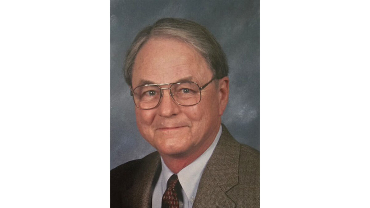 Portrait of an older man with glasses and gray hair, wearing a suit and tie, smiling against a blurred background.