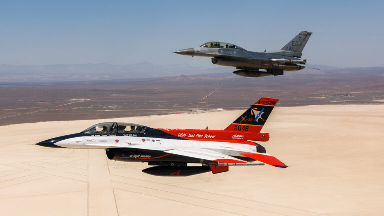 Two military jets flying in formation over a desert landscape, featuring one jet with a white, red, and black design and another in a gray camouflage pattern.