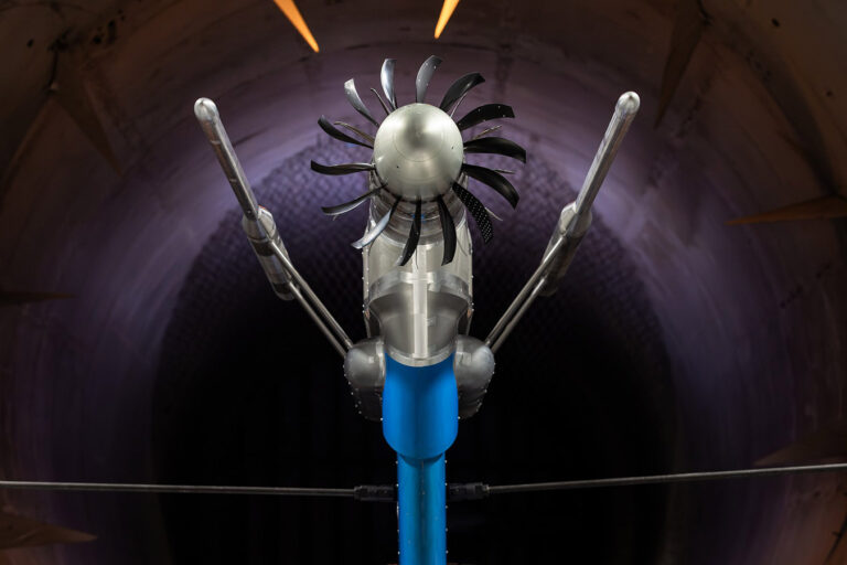 Close-up of a futuristic turbine prototype with several blades, mounted on a blue pole, inside a wind tunnel.