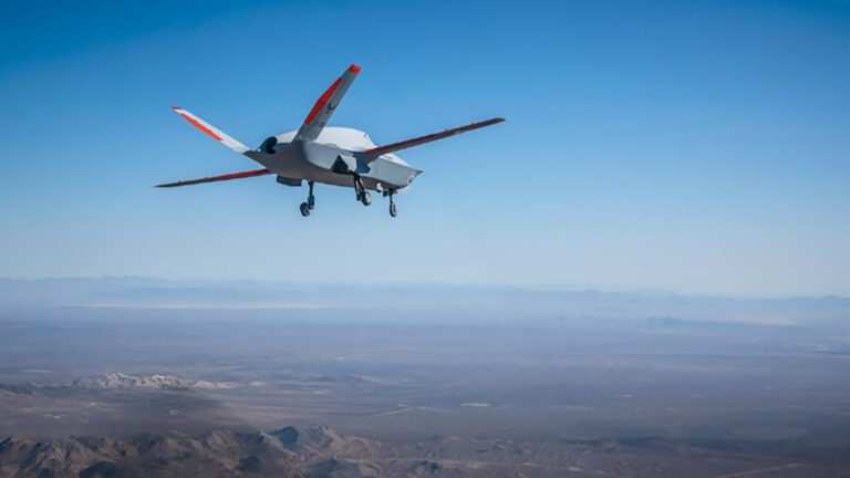 An unmanned aerial vehicle (UAV) flying over a vast desert landscape under a clear blue sky.