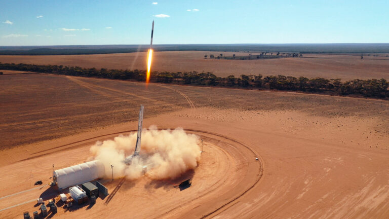 A rocket launches from a desert area, leaving a trail of smoke and dust. The landscape is flat with sparse vegetation and a clear sky.