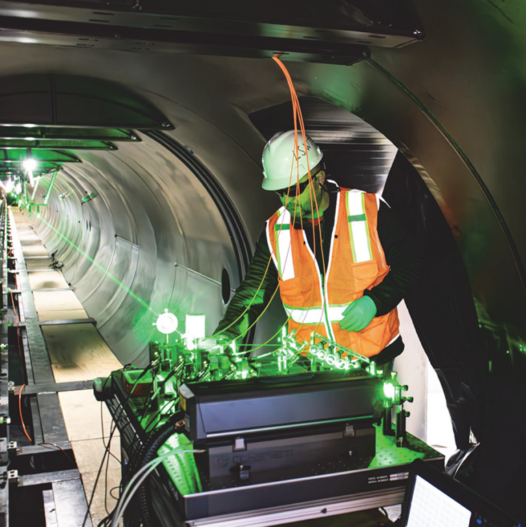 Worker in orange vest and hard hat operates equipment with green lights inside a metallic tunnel.
