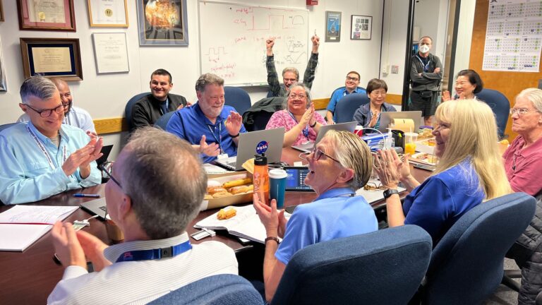 A group of people sit around a conference table, smiling and applauding. Some have laptops and notebooks. A person stands at the back, arms raised in celebration.