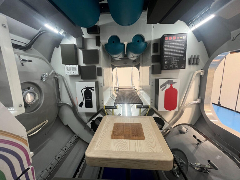 Interior view of a capsule or spacecraft module with control panels, fire extinguishers, and storage areas. The floor extends toward an open hatch.
