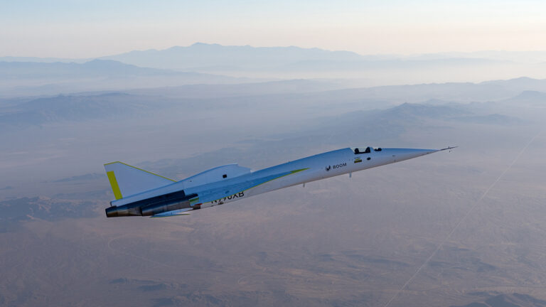 A white and blue supersonic jet flies over a vast desert landscape with mountains in the background under a clear sky.