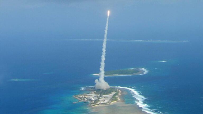 A rocket launches from a small island, leaving a trail of smoke against a clear blue sky, surrounded by ocean.