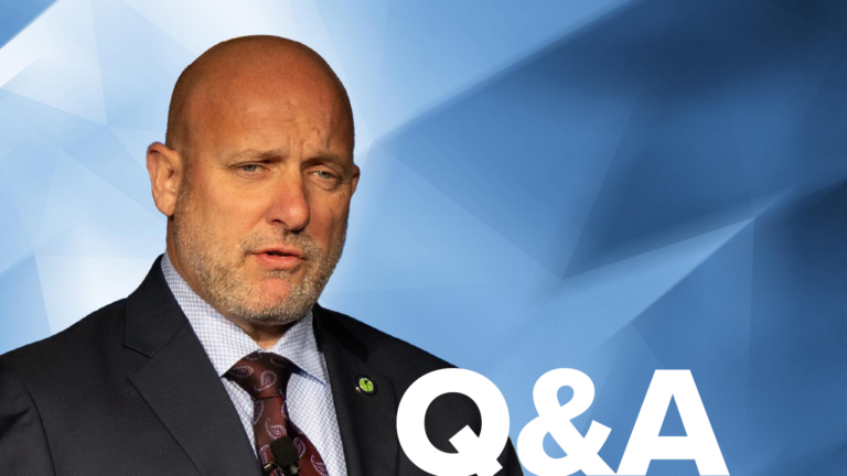 A bald man in a suit speaks in front of a geometric blue background with "Q&A" displayed in the lower right corner.