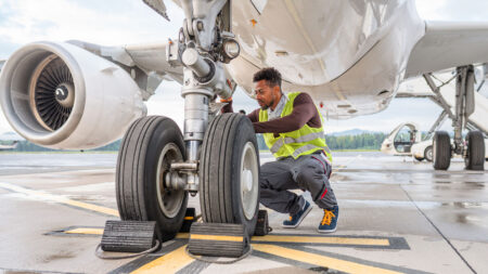 A maintenance worker in a high-visibility vest inspects the landing gear of a parked airplane on the tarmac.