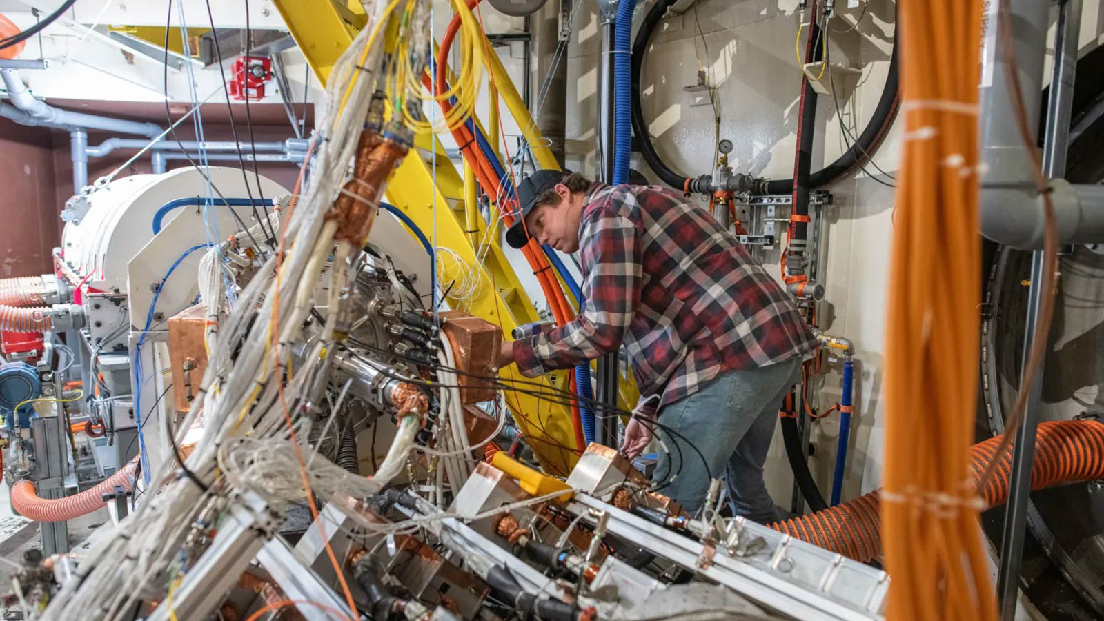 A person in a plaid shirt works on a complex arrangement of industrial machinery with numerous wires and pipes.