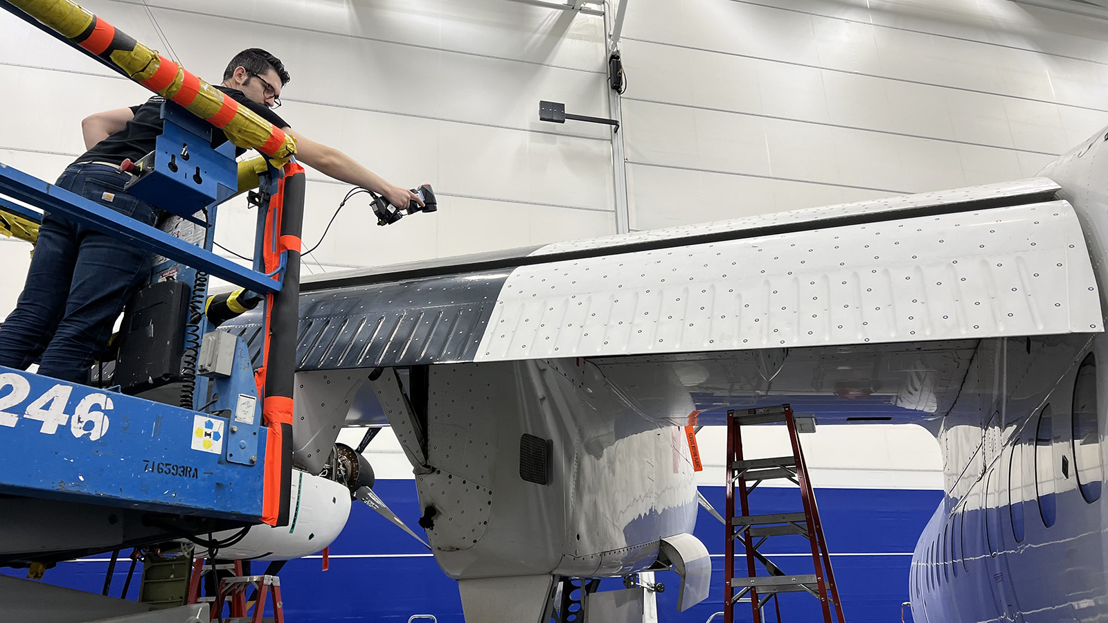 A person on a lift inspects an aircraft wing in a hangar.
