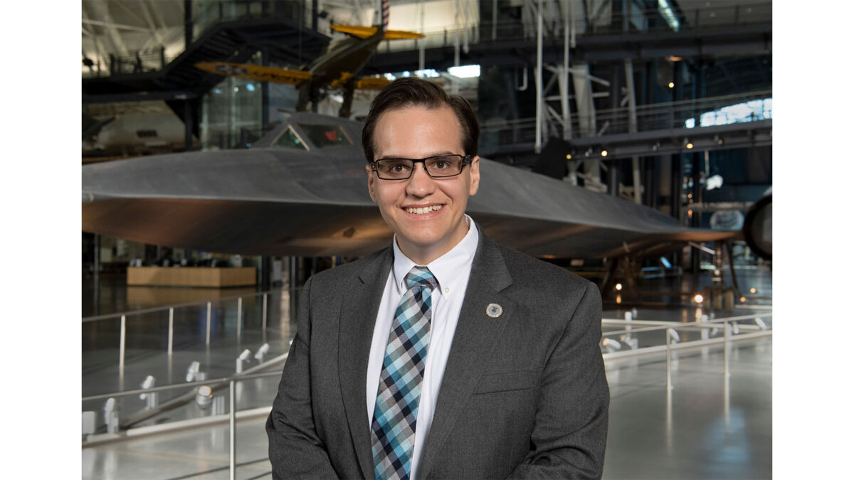 A person in a suit and tie stands in a museum, with an aircraft displayed in the background.