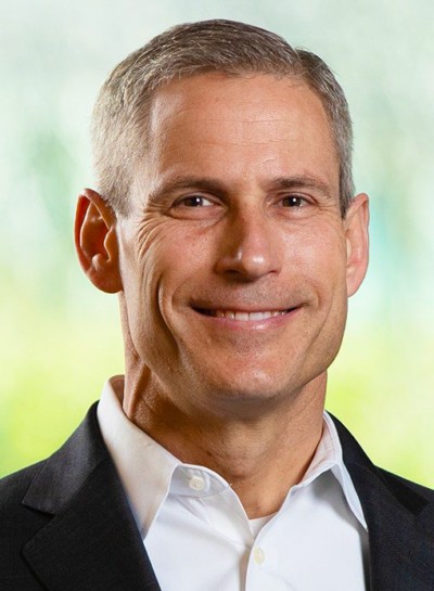 A person with short gray hair, wearing a suit and white shirt, smiling against a blurred background.