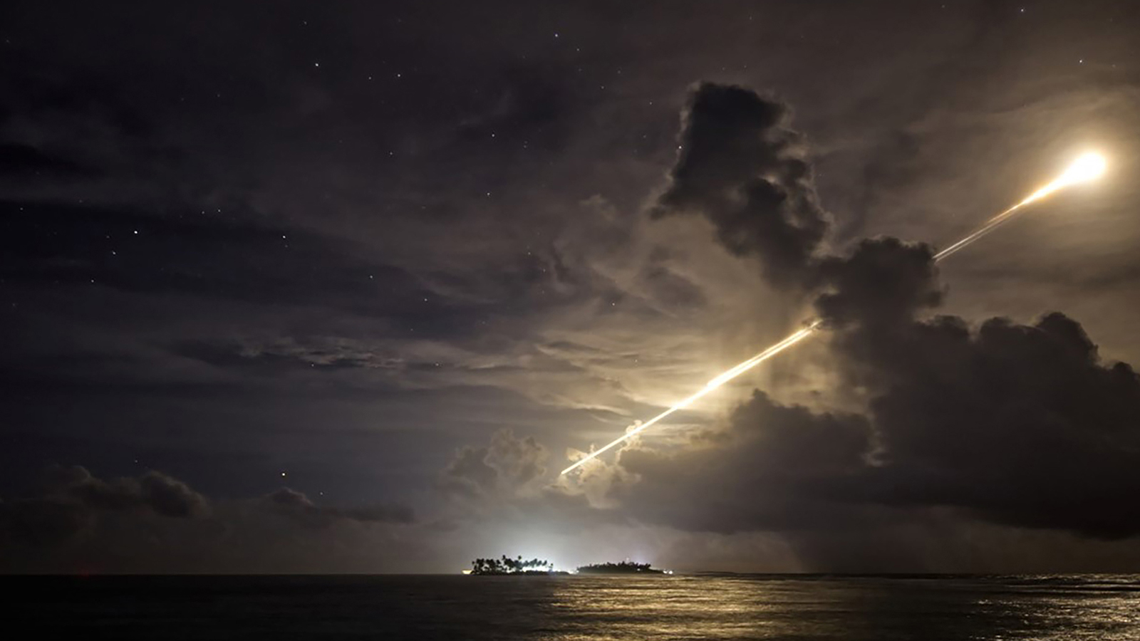 A bright light trails across a cloudy night sky above the ocean, with a small illuminated island in the distance.