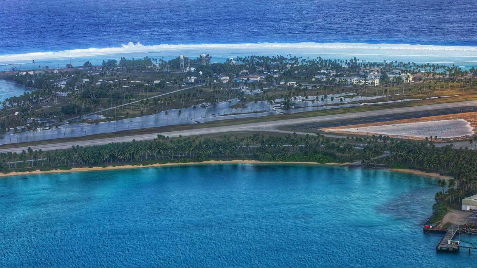 Aerial view of an atoll with a narrow airstrip, surrounded by turquoise and deep blue ocean waters, lined with palm trees and small buildings.