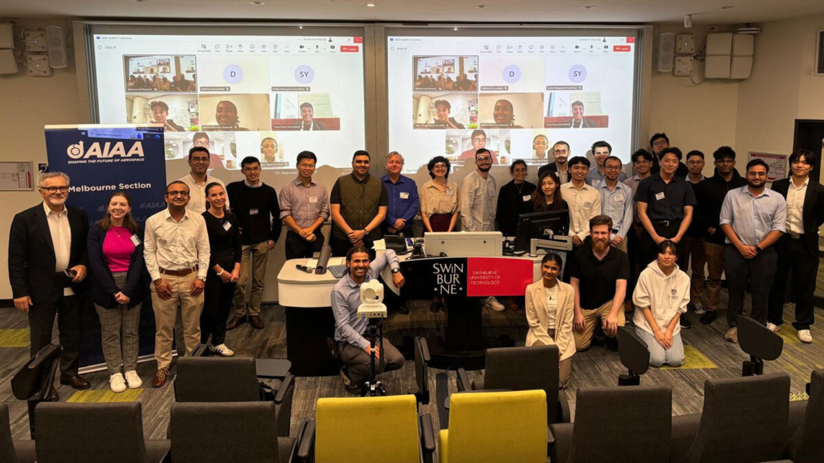 A group photo of around 30 people in a lecture room with Swinburne and AIAA banners. Some participants are posing near tables with screens displaying a video call.