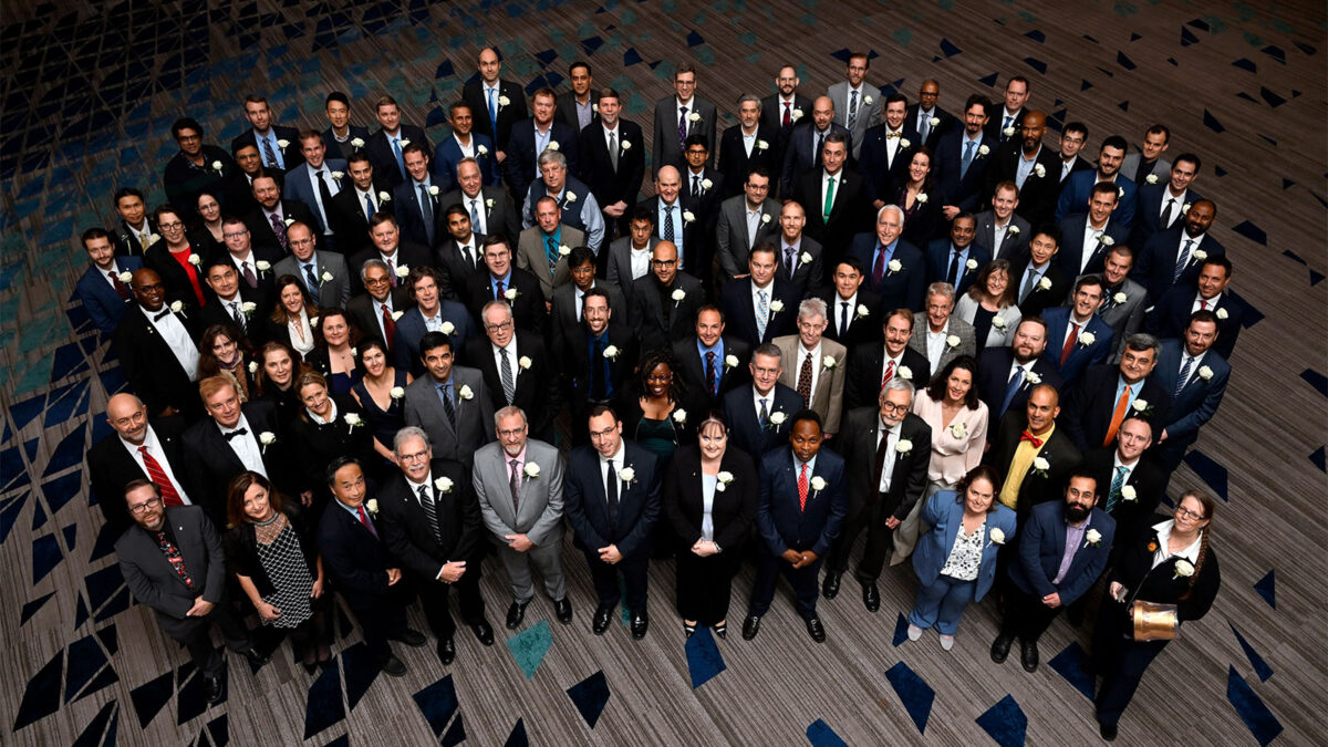 A large group of formally dressed people, standing closely together on a patterned floor, facing the camera. Many are wearing suits and dresses with corsages.