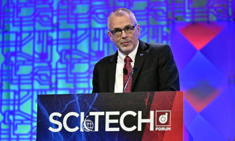 A man in a suit and tie speaks at a podium labeled "SciTech Forum" against a geometric patterned backdrop.