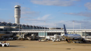 Airplane parked at an airport gate with a control tower and terminal building in the background under a partly cloudy sky.