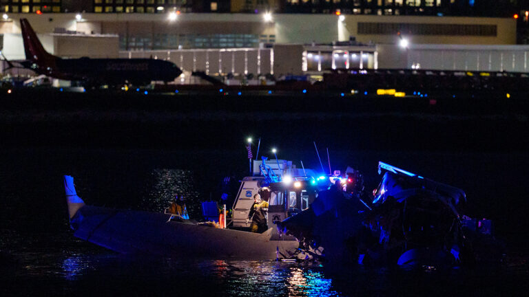 A boat with flashing lights approaches a partially submerged airplane at night near an airport, with a terminal and parked airplanes visible in the background.