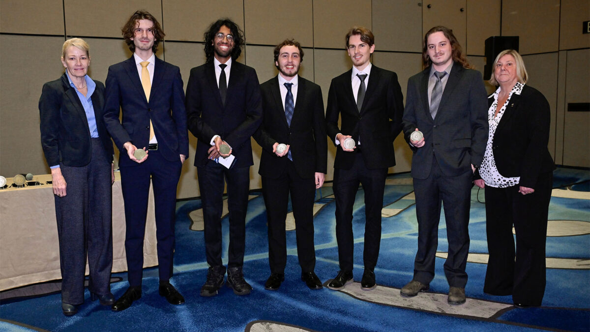 Seven people, five men in suits holding awards, and two women, stand in a row on a patterned blue carpet at an indoor event.