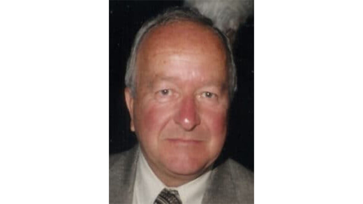 Elderly man in a suit and tie, with a neutral expression, against a dark background.