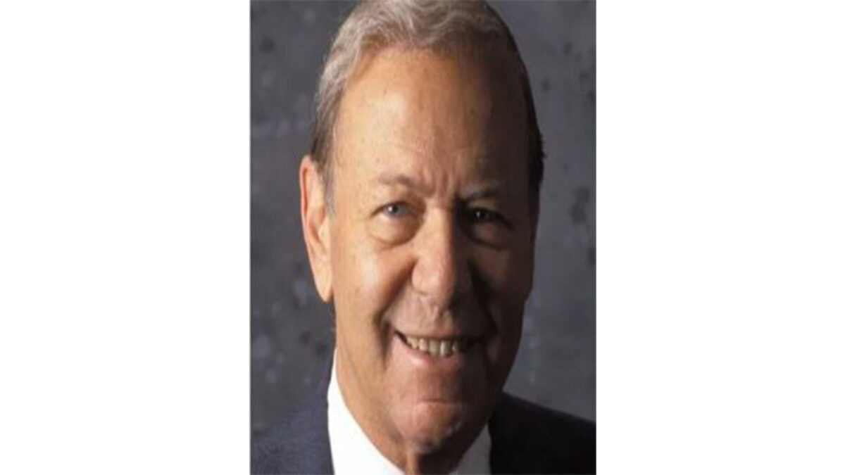 Portrait of an older man smiling, wearing a suit, with gray hair and a neutral gray background.