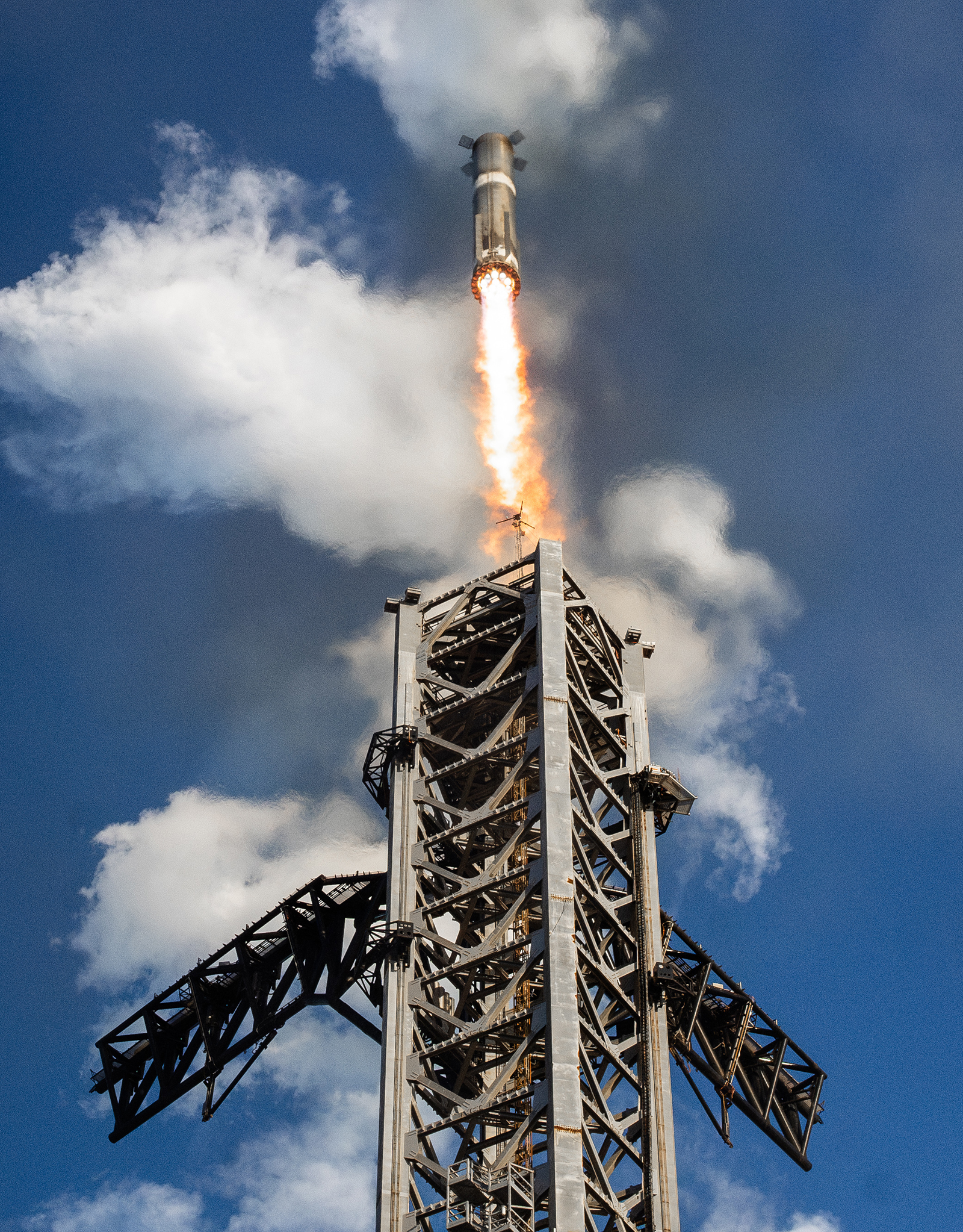 A rocket launches vertically into a blue sky with scattered clouds, emitting bright flames and smoke from its engines. A metal launch tower is visible in the foreground.