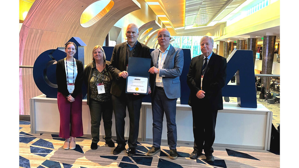 Five people stand indoors, one holding a certificate. Large stylized letters spell 