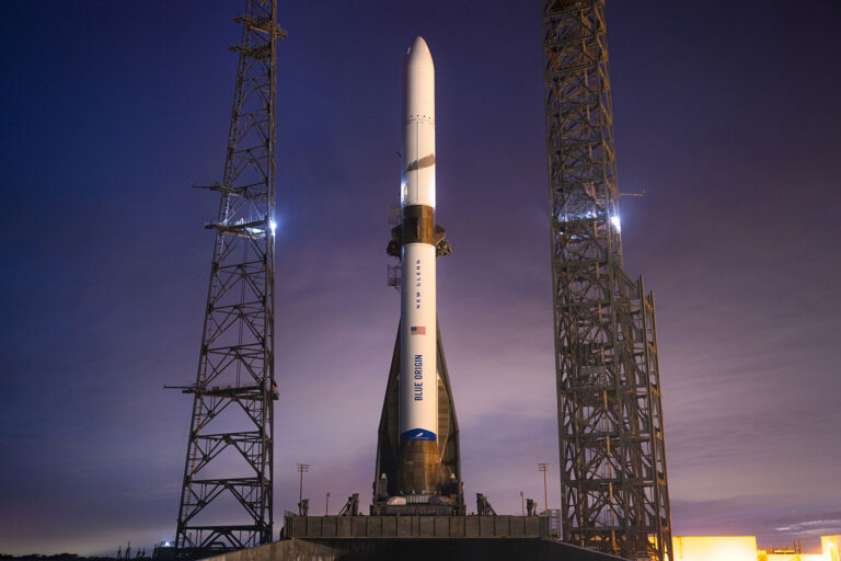 A rocket stands upright on a launch pad between two towers, with a blue sky in the background.